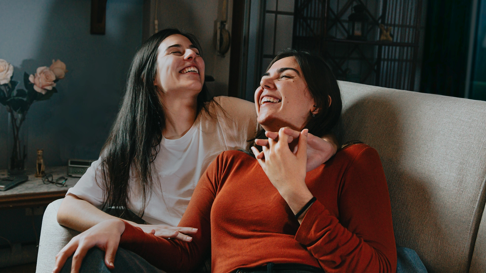 Two young women sitting on a couch laughing and holding each other's hands in a cozy room.